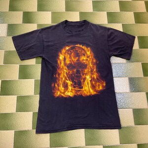 May include: Dark navy blue t-shirt with a flaming skull design. The skull is rendered in orange and yellow hues, with the word "OIL" above it. The shirt has short sleeves and a crew neck.