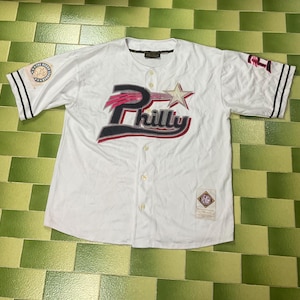 Peut inclure: Maillot de baseball blanc avec le mot "Philly" en rouge, noir et gris, avec une étoile. Le maillot a des rayures noires sur les manches et un petit écusson sur la manche. Le maillot est boutonné.