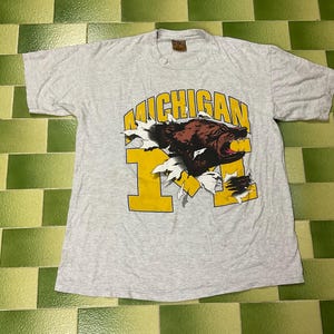 May include: A light grey short-sleeve t-shirt featuring a graphic of a brown bear head emerging from a tear in a large yellow letter M. The word MICHIGAN is printed in yellow above the bear. The shirt has a crew neckline and a tag at the collar.