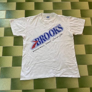 Puede incluir: Camiseta blanca con el logo azul y rojo de Brooks, con el texto "Basketball Shoes". La camiseta tiene cuello redondo y mangas cortas. La camiseta está tendida sobre una superficie de baldosas verdes y blancas.