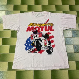 May include: White t-shirt featuring a motorcycle racing graphic. The design includes a motorcycle with a rider, the text "SCHWANTZ MOTUL", and the words "WORLD CHAMPION 500cc 1993". The graphic incorporates red, white, and blue colors.