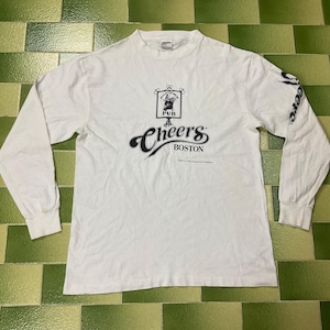 May include: White long-sleeve shirt with the text "Cheers Boston" in black script. The shirt features a pub logo above the text and "Cheers" printed on the sleeve. The shirt is made of a soft material.