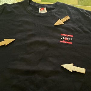 Może przedstawiać: Czarny t-shirt z małą metką Nike przy dekolcie. Koszulka posiada czerwono-białą grafikę z napisem "AUTHENTIC JORDAN BY NIKE". Koszulka wykonana jest z miękkiego materiału.
