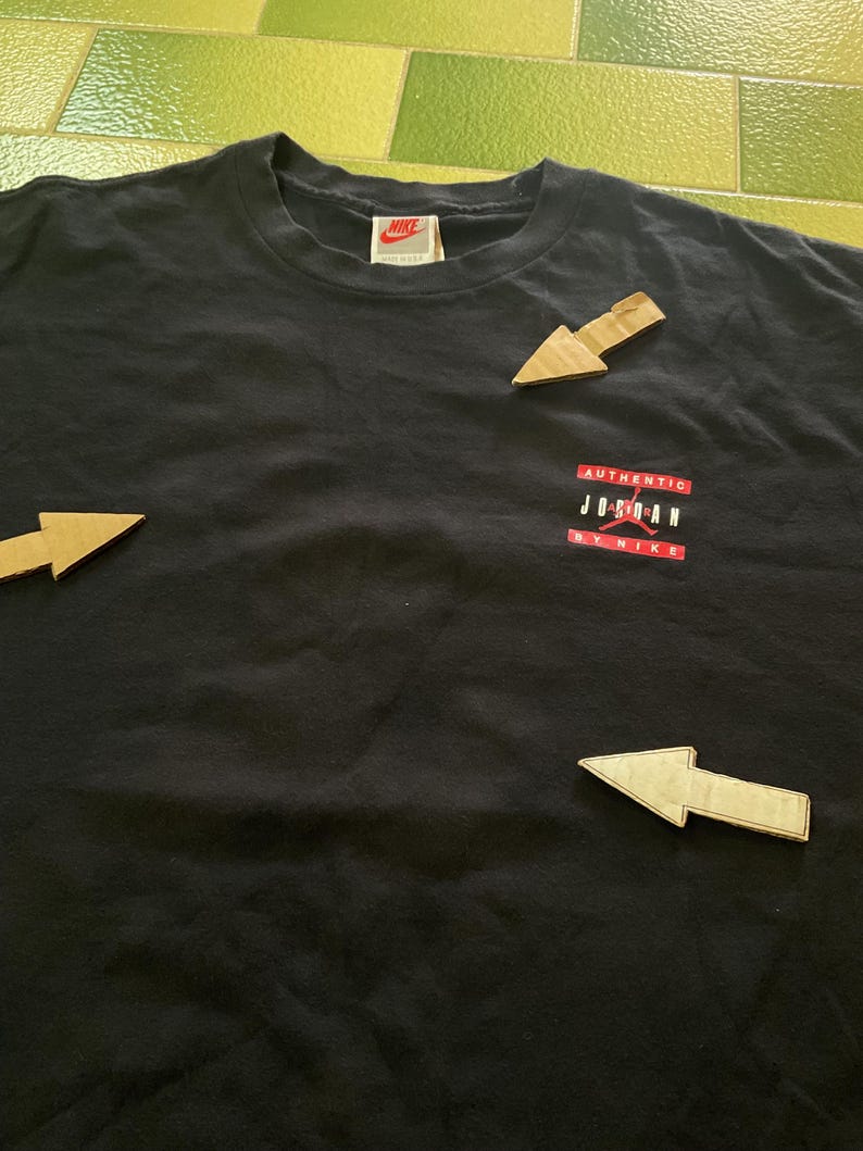 Może przedstawiać: Czarny t-shirt z małą metką Nike przy dekolcie. Koszulka posiada czerwono-białą grafikę z napisem "AUTHENTIC JORDAN BY NIKE". Koszulka wykonana jest z miękkiego materiału.