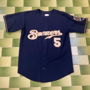 May include: Navy blue baseball jersey with the word "Brewers" in cream-colored script across the chest, along with the number "5". The jersey has cream-colored trim on the sleeves and collar. A team logo is on the left sleeve.