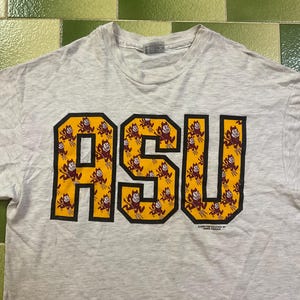Puede incluir: Camiseta gris claro con las letras "ASU" en letras grandes, en negrita, amarillas y granates. Las letras están delineadas en negro. Las letras están rellenas con un patrón repetido de una mascota de diablo granate.