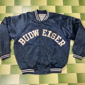 May include: Navy blue satin bomber jacket with the text "BUDW EISER" in white block letters across the chest. The jacket has a striped collar, cuffs, and hem. A vintage style jacket.
