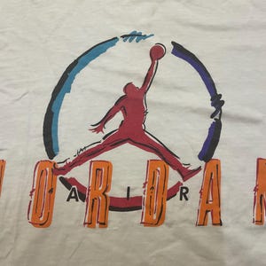 Puede incluir: Camiseta blanca con el logo de Jordan. El logo muestra una silueta roja de un jugador de baloncesto saltando por un bal&oacute;n. La palabra "JORDAN" est&aacute; escrita en letras naranjas debajo.