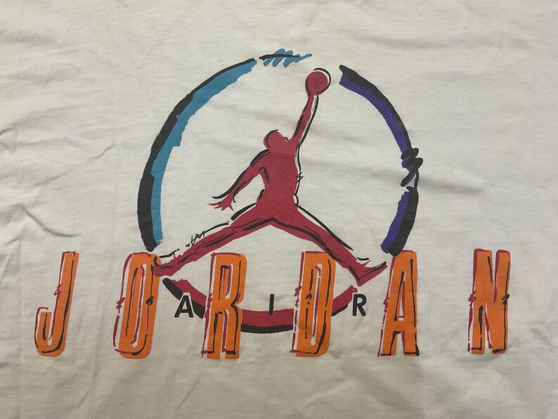 Puede incluir: Camiseta blanca con el logo de Jordan. El logo muestra una silueta roja de un jugador de baloncesto saltando por un bal&oacute;n. La palabra "JORDAN" est&aacute; escrita en letras naranjas debajo.
