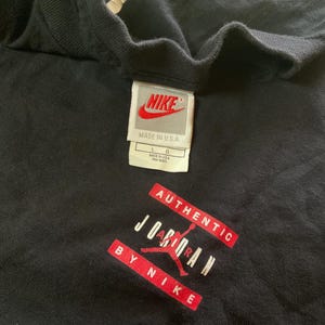 Może przedstawiać: Czarny t-shirt z metką Nike i napisem "AUTHENTIC JORDAN BY NIKE" w kolorze czerwono-białym. Na metce napis "NIKE MADE IN U.S.A." i rozmiar L. Koszulka wykonana jest z miękkiego materiału.
