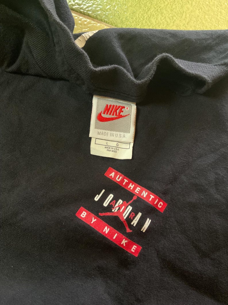 Może przedstawiać: Czarny t-shirt z metką Nike i napisem "AUTHENTIC JORDAN BY NIKE" w kolorze czerwono-białym. Na metce napis "NIKE MADE IN U.S.A." i rozmiar L. Koszulka wykonana jest z miękkiego materiału.