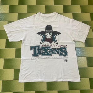 May include: White t-shirt featuring a graphic of a cowboy with a star and the text "SAN ANTONIO TEXANS". The shirt also has the text "PROFESSIONAL FOOTBALL".