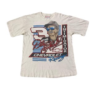 May include: Off-white t-shirt featuring a portrait of a man wearing sunglasses and a hat, with the number 3 and the words "Intimidator" and "Chevrolet Racing". The shirt has a vintage aesthetic.