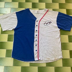 May include: A gray and blue baseball jersey with a button-up front and short sleeves. The jersey has a red stripe along the button line and a logo on the chest. The sleeves are blue, and the jersey is made of a soft material.