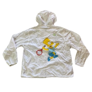 May include: White hooded jacket featuring a cartoon image of Bart Simpson playing tennis. The graphic shows Bart in a blue shirt and shoes, holding a red tennis racket. Text on the jacket reads "SERVING UP TROUBLE, MAN!".
