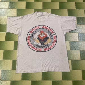 Puede incluir: Camiseta blanca con un gráfico circular que presenta un león de dibujos animados haciendo una señal de aprobación. El gráfico incluye el texto "DRUG ABUSE POLICE EDUCATION" y "RESISTANCE SCHOOL PARENTS".