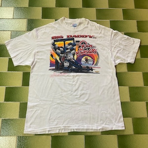May include: White t-shirt featuring a colorful graphic of a race car with the text "Big Daddy" and "Don Garlits". The design includes a checkered flag and the words "Grand Bay, AL".