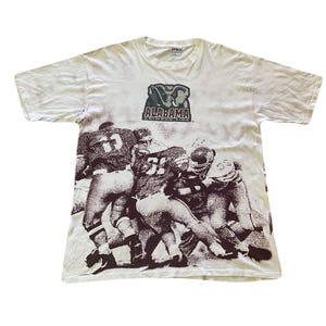 May include: A white short-sleeve t-shirt featuring a dark brown graphic print of a football game in progress. The top center displays an elephant logo with the text ALABAMA CRIMSON TIDE. The shirt has a crew neckline and a standard fit.