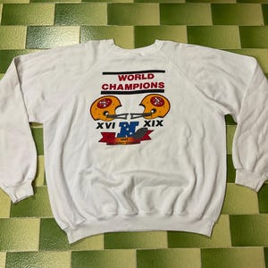 May include: A white long-sleeve sweatshirt featuring a graphic print that reads WORLD CHAMPIONS with two gold football helmets, Roman numerals XVI and XIX, a blue letter N, and a red banner with the text 1987 THE WEST CHANCE printed below.