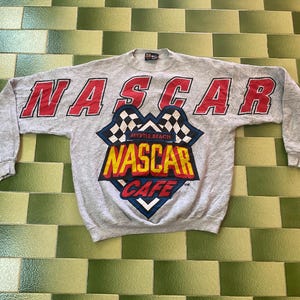 May include: A gray sweatshirt with the word "NASCAR" in red block letters across the chest and sleeves. Below the word "NASCAR" is a graphic with a checkered flag and the words "MYRTLE BEACH" and "NASCAR CAFE" in yellow and red.