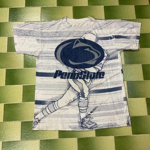 Puede incluir: Camiseta blanca con un gráfico de un jugador de fútbol americano y el logo de Penn State en azul marino. La camiseta tiene un estampado de rayas en tonos blancos y azules.