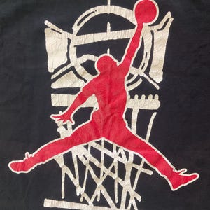 Może przedstawiać: Czarna koszulka z czerwoną sylwetką koszykarza w trakcie wsadu. Zawodnik znajduje się przed białym koszem do koszyk&oacute;wki i tablicą. Projekt ma postarzany, vintage wygląd.