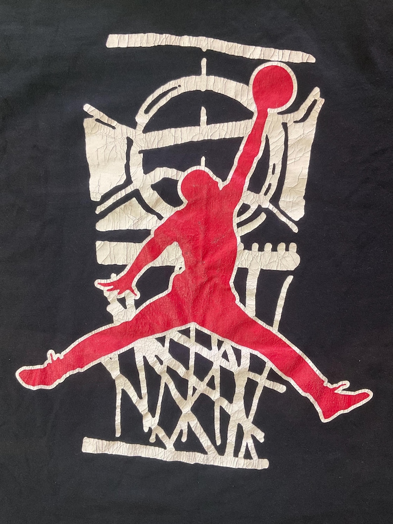 Może przedstawiać: Czarna koszulka z czerwoną sylwetką koszykarza w trakcie wsadu. Zawodnik znajduje się przed białym koszem do koszyk&oacute;wki i tablicą. Projekt ma postarzany, vintage wygląd.