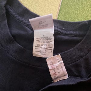 Może przedstawiać: Zbliżenie czarnej koszulki z dwoma białymi metkami. Etykiety zawierają logo Nike, informacje o rozmiarze i instrukcje pielęgnacji. Koszulka jest w rozmiarze L, wyprodukowana w USA i jest w 100% bawełniana.