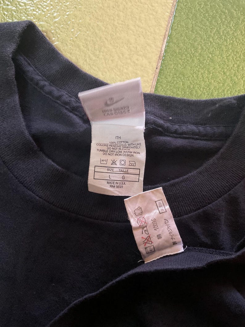 Może przedstawiać: Zbliżenie czarnej koszulki z dwoma białymi metkami. Etykiety zawierają logo Nike, informacje o rozmiarze i instrukcje pielęgnacji. Koszulka jest w rozmiarze L, wyprodukowana w USA i jest w 100% bawełniana.