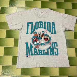 May include: A light gray t-shirt featuring the text "FLORIDA MARLINS" in teal. A baseball graphic with cartoon characters is in the center. The MLB logo is at the bottom. The shirt is made of a soft material.