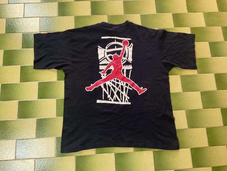 Może przedstawiać: Czarny t-shirt z czerwoną grafiką koszykarza w powietrzu, zaraz po wsadzie piłki. Grafika umieszczona na białym tle z motywem kosza do koszyk&oacute;wki. Koszulka w klasycznym stylu sportowym.