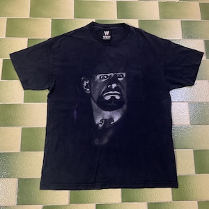 May include: Black t-shirt featuring a grayscale image of a man's face. The image is centered on the shirt, with the man's face prominently displayed. The shirt has a crew neck and short sleeves. The WWE logo is visible on the collar.