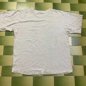 Puede incluir: Una camiseta blanca lisa de manga corta, colocada sobre una superficie de baldosas verdes y blancas. La camiseta parece estar hecha de una tela suave y ligera. La camiseta est&aacute; ligeramente arrugada.