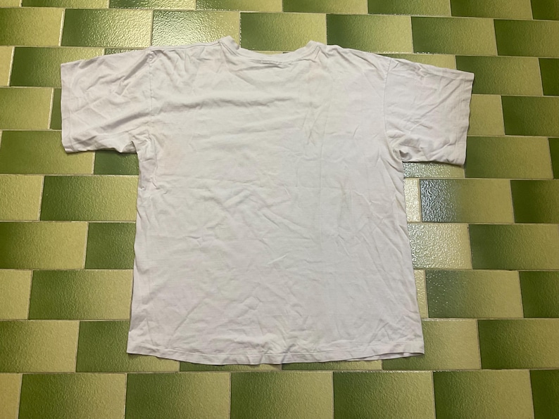 Puede incluir: Una camiseta blanca lisa de manga corta, colocada sobre una superficie de baldosas verdes y blancas. La camiseta parece estar hecha de una tela suave y ligera. La camiseta est&aacute; ligeramente arrugada.