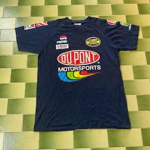 May include: Navy blue short-sleeved t-shirt featuring the "DuPont Motorsports" logo in red and white, with a colorful arc design. The shirt also displays "Pepsi", "GMAC", and "NASCAR Nextel Cup Series" logos.