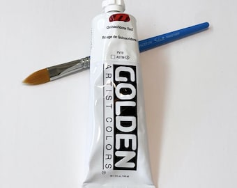 May include: A white tube of Golden Artist Colors Quinacridone Red paint with black text and a red square logo, positioned vertically. A blue-handled paintbrush with a brown bristle tip is placed horizontally across the paint tube.