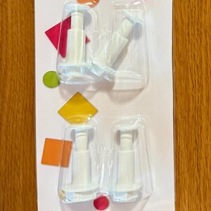 May include: A package of Sweetshop pop-out cutters for round and square shapes. The package includes four cutters, with the text "Make Something Remarkable" at the top. The package has colorful shapes and the text "COUPEURS À SORTIR ROND ET CARRÉ".