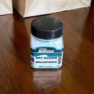 May include: A small, clear jar with a black lid, labeled "DRY MEDIUM WOLLASTONITE" and "50ml". The jar contains a white substance. The label has blue and white accents. The jar is on a wooden surface.