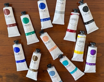 May include: A collection of Gamblin artist oil paint tubes in various colors, including red, green, blue, and yellow. The tubes are arranged on a dark wooden surface, showcasing the vibrant pigments.