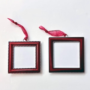 May include: Two square red picture frames with silver trim and white centers, each suspended by a red ribbon. The frames have a textured border and are set against a white background. Ideal for displaying small photos or artwork.