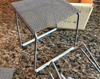 May include: A square, grey metal mesh platform supported by four thin, light grey metal legs. The structure is modular, with additional detached metal leg components and a separate piece of grey mesh visible nearby on the surface.