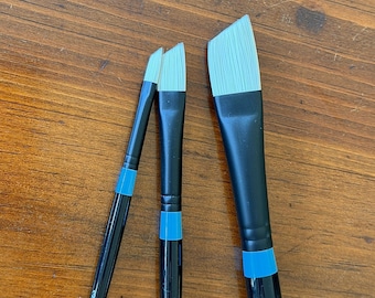 May include: Three Princeton Aspen paint brushes with black handles, bright blue ferrules, and white synthetic bristles. The brushes feature angled, chisel-edge tips of varying widths, with the text "ASPEN" and "PRINCETON" printed on the handles.