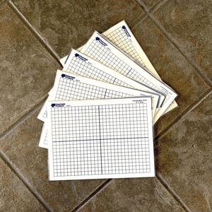 May include: A stack of white rectangular plastic sheets featuring a black printed centimeter grid with an X-Y axis. Each sheet is enclosed in a clear protective sleeve and displays the Classroom Products logo and the text Centimeter Grid X-Y Axis.