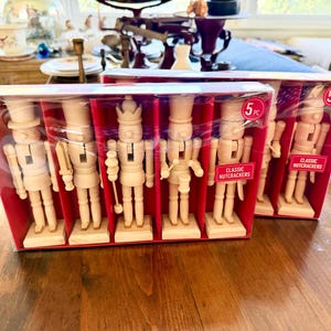 May include: Two sets of five unfinished wooden nutcracker figurines in clear plastic packaging. Each set contains five distinct figures with varied hats and accessories. The packaging features red cardboard inserts and pink labels that read 5 PC and CLASSIC NUTCRACKERS.