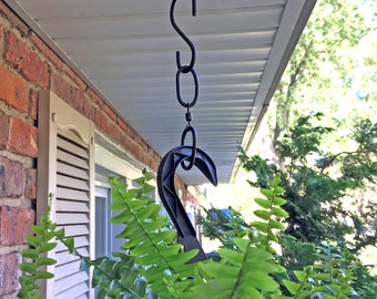 May include: A black hanging plant bracket with an S-hook, chain, and curved hook, suspended from a white soffit. Lush green fern fronds fill the foreground, with a brick wall and window shutters in the background.
