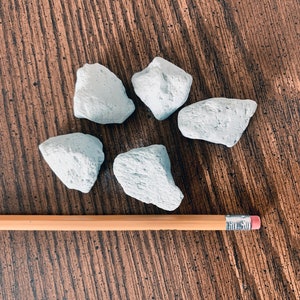 May include: Five light blue, irregularly shaped rocks are arranged on a brown wooden surface. A wooden pencil with a pink eraser is placed next to the rocks for scale. The rocks appear to be natural and textured.