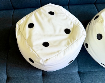 May include: A white, cube-shaped ottoman designed to resemble a six-sided die. The soft, fabric-covered piece features five distinct black circular spots arranged on its top and side surfaces, mimicking the pips found on a standard game die.