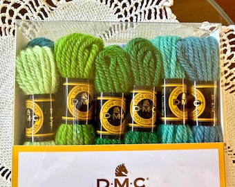 May include: A set of six skeins of DMC Laine Colbert tapestry wool in a gradient of green and blue shades, packaged in a clear plastic box. Each skein features a black and yellow label with the DMC logo and text indicating 8m - 8.7yd length.