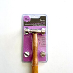 May include: A jewelry-making texture hammer with removable heads, packaged in a clear plastic container. The hammer has a wooden handle and a silver-colored metal head. The package includes two interchangeable textured heads, one with a striped pattern and one with a dotted pattern.