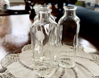 May include: Three clear glass bottles with a rectangular shape are arranged on a white lace doily. The bottles have a small, square base and a narrow neck. The bottles are empty and appear to be vintage.